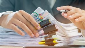 Close-up of hands sorting through stacks of documents clipped together with colorful paper clips and binder clips while holding a pen